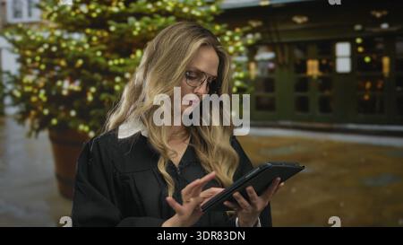 Young blonde woman in a judge uniform uses a tablet outdoors with a festive backdrop featuring a decorated christmas tree. Stock Photo