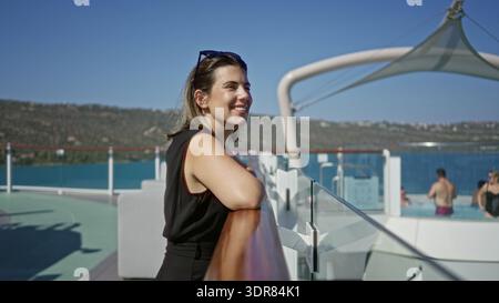 Woman leaning on ship rail with arm resting on wooden railing on deck building, smiling toward the ocean horizon with sunglasses on head; serenity. Stock Photo