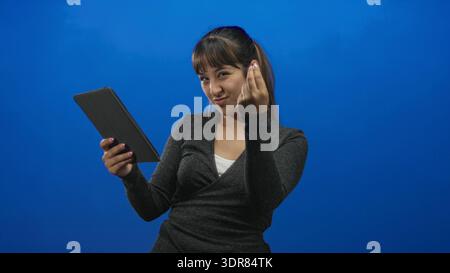 Woman holding tablet makes finger heart gesture and smiles in studio with blue backdrop; playful digital connection. Stock Photo