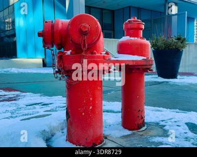 A red fire hydrant stands on a city sidewalk with snow around it during winter. The cold weather creates a strong contrast between the hydrant and the Stock Photo