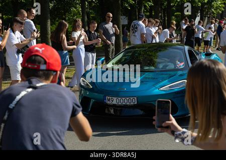Blue McLaren sports car is showcased as a crowd of spectators takes photos during a car show event, with trees lining the background in a public area Stock Photo