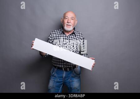 Older Man Holds Blank Sign in Front of Gray Background While Smiling During Indoor Session Stock Photo
