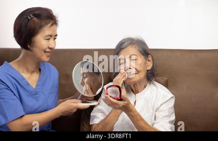 Elderly woman applying face powder with care giver in nursing home. Stock Photo