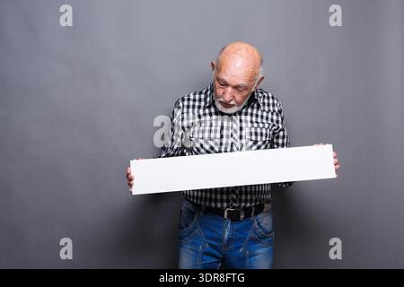 Older Man Holds Blank Sign While Looking Down in Studio With Gray Background Stock Photo