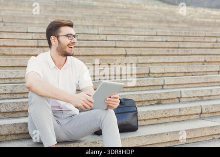 Young Man Sits on Steps Smiling While Using Tablet Outdoors in the City During the Day Stock Photo