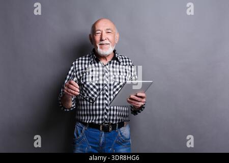 Senior Man Holds Tablet and Smiles While Standing Against Gray Background in a Casual Setting Stock Photo