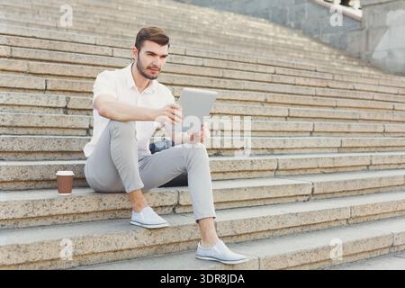Young Man Sits on Stairs With Tablet While Drinking Coffee Outdoors in a Public Area During the Day Stock Photo