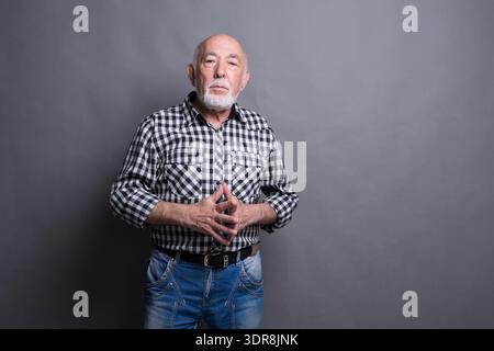 Man Stands With Hands Clasped in Front of Gray Background While Wearing Checkered Shirt and Jeans Stock Photo