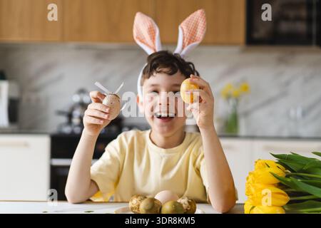 Smiling child wearing bunny ears with natural Easter eggs at home kitchen Stock Photo