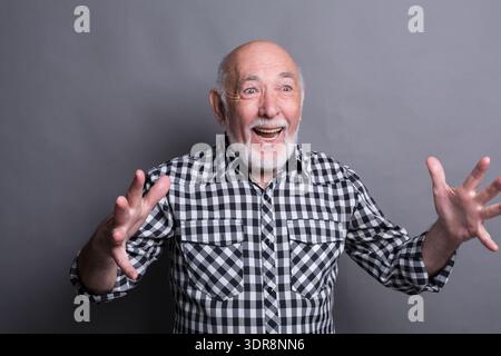 Elderly Man Smiles and Gestures With Hands Against Gray Background Stock Photo