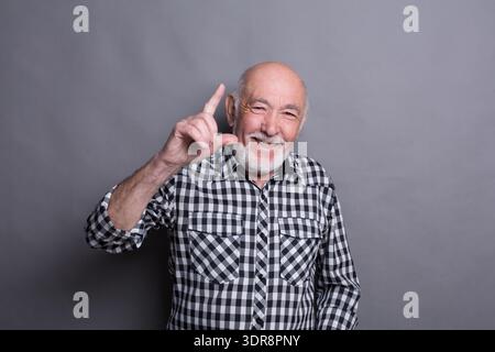 Elderly Man With Gray Beard Smiles and Shows Hand Gesture During Indoor Activity Stock Photo