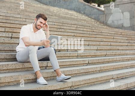 Man Sits on Stairs and Uses Tablet While Smiling During the Day in an Urban Area Stock Photo