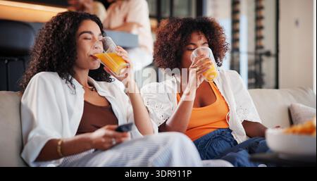 Women, friends and watching tv with drinks on sofa with juice, bonding and relax in home for movies. People, glasses and television on couch for Stock Photo
