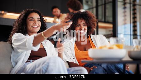 Women, friends and watching tv on sofa with smile, remote control and movie choice in living room. People, happy and relax on couch with streaming Stock Photo