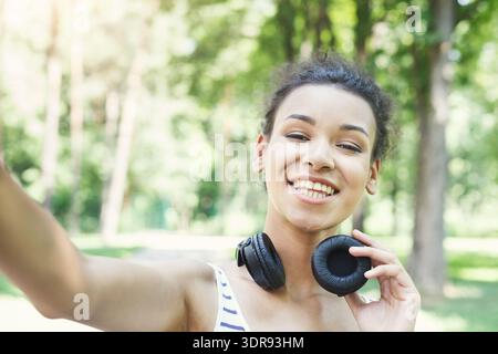 Young Woman Smiling and Taking a Selfie While Wearing Headphones in a Park Setting Stock Photo