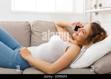 Young Woman Talks on the Phone While Lying on the Couch in a Bright Living Room Stock Photo