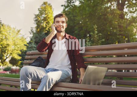 Young Man in Park Talking on Phone While Sitting on Bench With Laptop Present Stock Photo