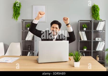 Happy excited man working on laptop in office and making yes gesture rejoicing in successful job Stock Photo