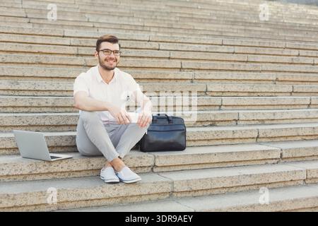 Man Sits on Steps With Laptop and Bag Enjoying Break in Urban Location Stock Photo