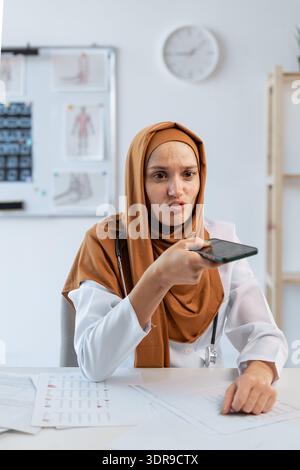 Muslim female doctor in hijab using smartphone for AI voice recognition in hospital or clinic. Stock Photo