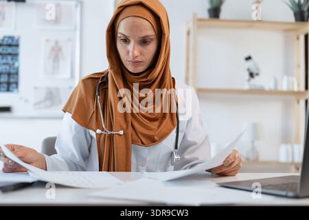 Muslim woman doctor with stethoscope reading medical documents and reports sitting at desk working with laptop computer in hospital or clinic Stock Photo