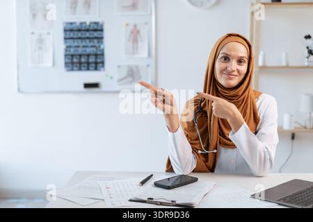 Smiling young muslim woman doctor wearing a hijab pointing with finger on space for advertisement while communicating and working in modern clinic Stock Photo