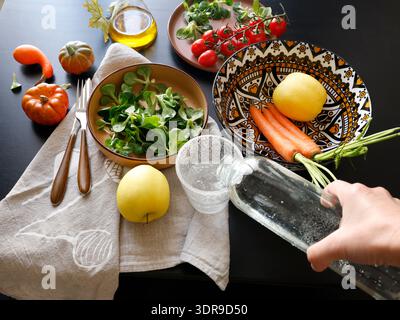 Anonymous person pours water from bottle into glass. Multicolored ceramic plates and patterned bowl with vegetables and herbs on black background Stock Photo