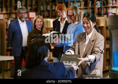 Young Asian Woman Handing Book to Caucasian Woman during Signing Event Stock Photo