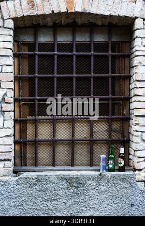 Three beverage containers including beer bottles and an energy drink can left abandoned on the stone sill of a rusty iron-barred basement window set i Stock Photo