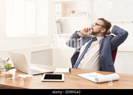 Business Professional Taking a Call While Sitting at Desk With Laptop and Tablet in Office Stock Photo