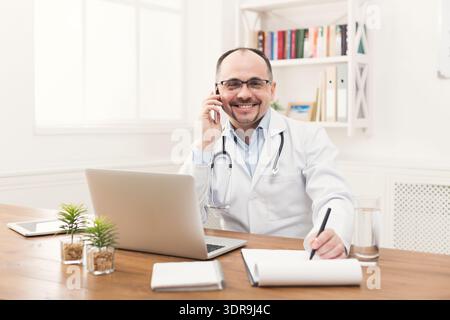 Doctor Talks on Phone While Taking Notes in Office During Working Hours Stock Photo