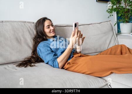 Smiling positive woman artist lying on couch scrolling social media online on smartphone at home. Stock Photo