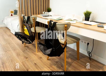 Desk stocked with books for homework and backpacks hanging on chairs in a cozy apartment. Organized study space ready for learning and education after Stock Photo