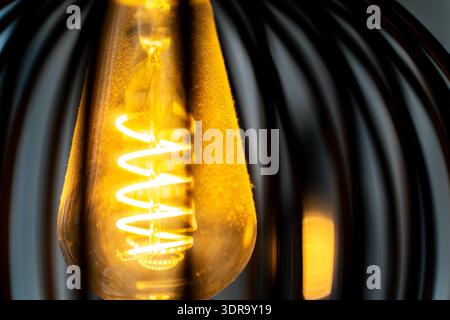 Spiral warm filament of a glowing bright light bulb inside a lampshade. The illuminated coil creates a cozy and inviting atmosphere with soft amber li Stock Photo