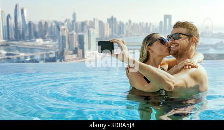romantic couple taking selfie in rooftop infinity swimming pool with Dubai skyline in background. banner with copy space Stock Photo