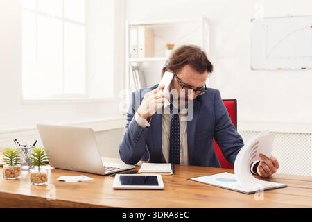 Businessman Working at Desk While Talking on Phone and Reviewing Documents Stock Photo