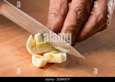 Chef in apron cuts the garlic with a knife. Closeup view of the hands of an adult woman cutting a garlic with a kitchen knife on a cutting board Stock Photo