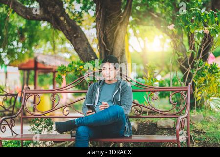 Handsome man sitting on a bench checking cell phone at sunset. Young man sitting on a park bench texting on cell phone Stock Photo