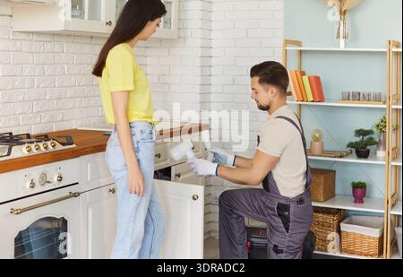 Man plumber in uniform cleaning siphon under kitchen sink, telling about problem to woman client Stock Photo