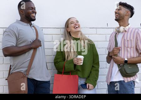 Diverse friends laughing and talking against painted white brick wall with coffee, bags, headphones Stock Photo