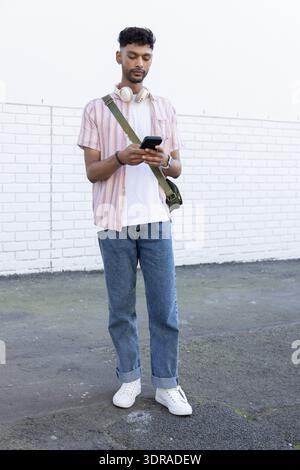 Indian man in twenties standing by white brick wall using smartphone with headphones, green bag Stock Photo