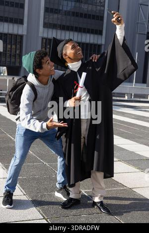 African American males holding phone taking selfie on tiled plaza wearing cap and holding diploma Stock Photo