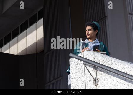 Teenage African American student leaning on railing holding phone and notebook on right, copy space Stock Photo