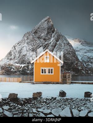 View of a vibrant yellow cabin nestled in a snowy landscape beneath a towering mountain peak, creating a striking contrast, Reine, Nordland, Norway. Stock Photo