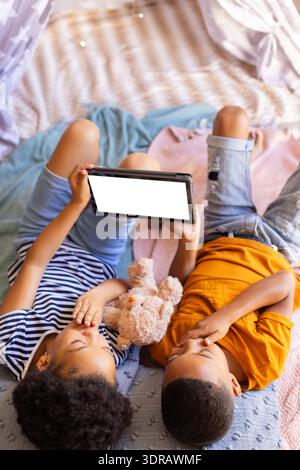 Brother and sister lying on bed using tablet and holding teddy bear at home, copy space, in teepee Stock Photo