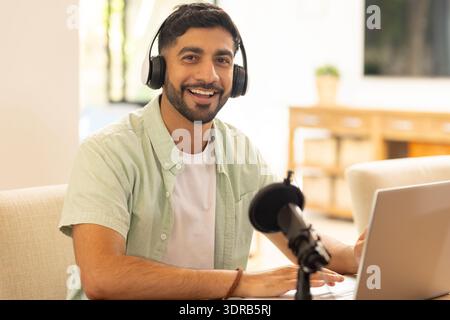 Smiling man using laptop and microphone, wearing headphones, working from home Stock Photo