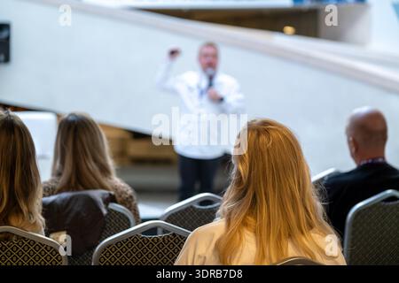 Speaker presents to seated audience at conference hall. Audience faces stage with microphone and projection area. Professional event atmosphere focused on talk and learning. Stock Photo