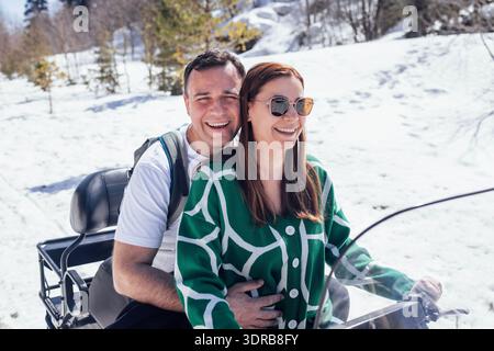 A young married couple is spending a vacation in the mountains. An attractive man and a woman are riding an ATV on a snowy road. Stock Photo
