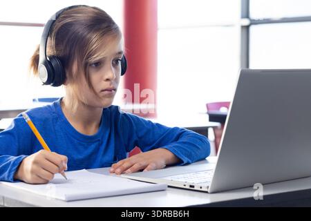 In school, boy wearing headphones writing in notebook while using laptop Stock Photo