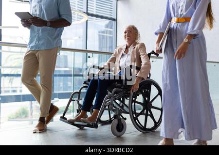 Discussing work, business professionals walking with woman in wheelchair in office hallway Stock Photo